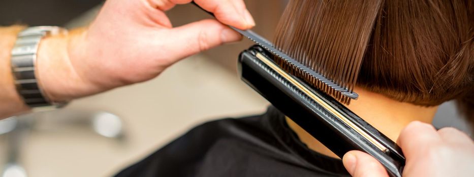 Close up of the hairdresser straightening the short hair of a female client with a hair straightening iron in a beauty salon. Selective focus