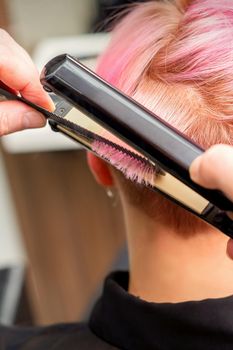 Close up of the hairdresser straightening the short pink hair of a female client with a hair straightening iron in a beauty salon