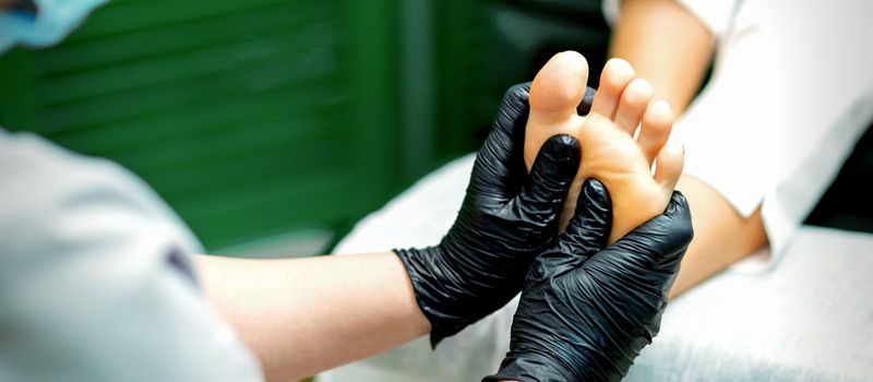 Beautician in protective rubber gloves doing massage on the sole of the female foot in a spa beauty salon