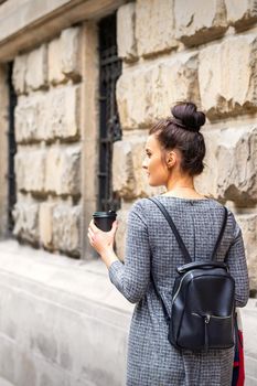 Back view of a young woman with a hot drink and backpack walks in a European city