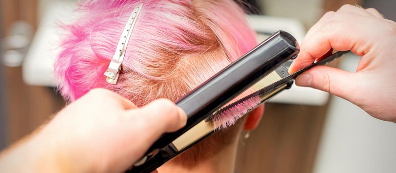 Close up of the hairdresser straightening the short pink hair of a female client with a hair straightening iron in a beauty salon