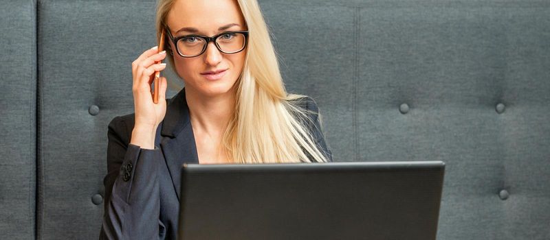 Portrait of a young caucasian businesswoman in glasses calls on a smartphone with a laptop at the table looking at the camera