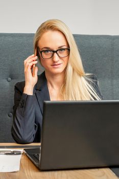 Portrait of a young caucasian businesswoman in glasses calls on a smartphone with a laptop at the table looking at the camera