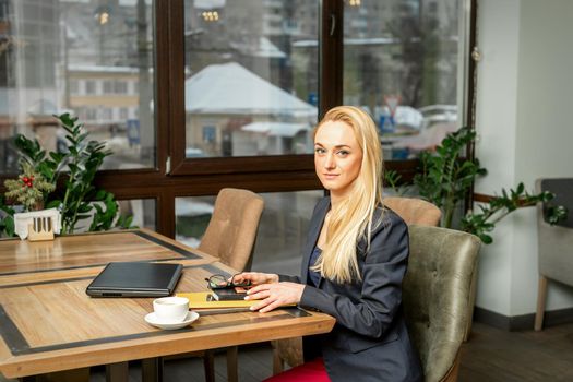 Portrait of a young caucasian businesswoman with documents and laptop at the table looking at the camera in cafe