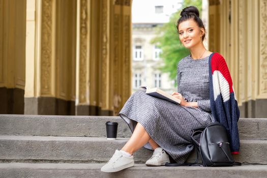 Portrait of a young female student with a book sitting on the stairs of building outdoors