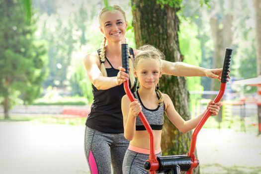 Mother with daughter in sportswear is doing sport exercises on a simulator in the summer park