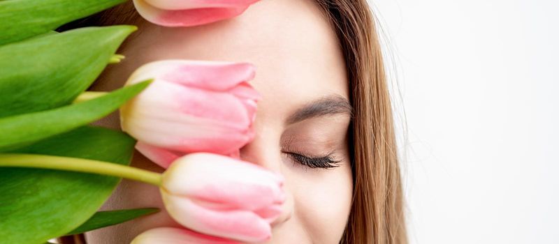 Half face portrait of a happy young caucasian woman with closed eyes and pink tulips cover her face against a white background