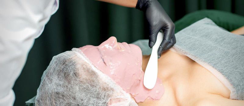 The cosmetologist applying an alginic mask to the face of a young woman in a beauty salon