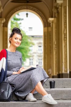 Portrait of a young female student with a book sitting on the stairs of building outdoors