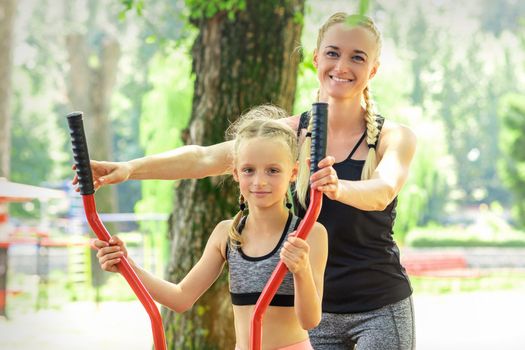 Mother with daughter in sportswear is doing sport exercises on a simulator in the summer park