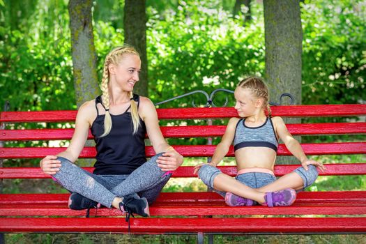 Mother with daughter wearing sportswear sitting on the bench in the summer park