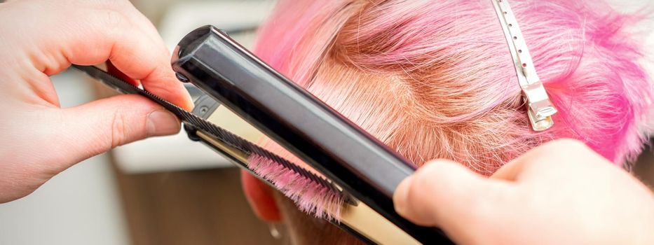 Close up of the hairdresser straightening the short pink hair of a female client with a hair straightening iron in a beauty salon