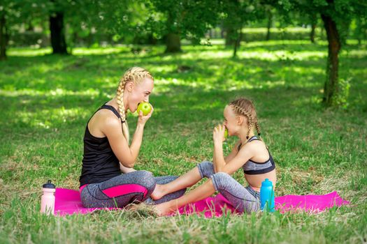 Mother with daughter in sportswear eats apples sitting on a mat in the summer park