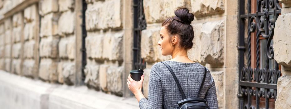 Back view of a young woman with a hot drink and backpack walks in a European city