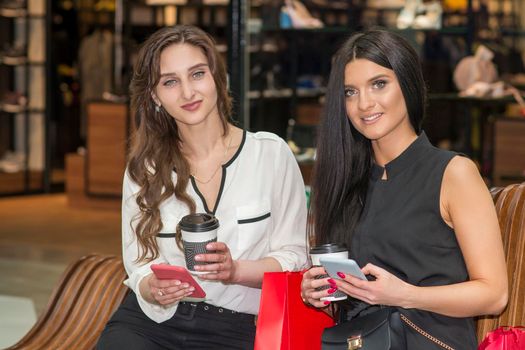 Two beautiful caucasian young women looking at the camera with bags sitting on the bench while resting in the shopping mall