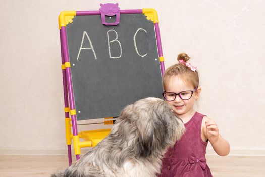 a little girl teacher in a dress and glasses sits on the floor near the blackboard and shows her dog. Pet does not want to learn and licks her mistress