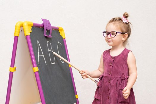 cheerful emotional girl of three years with a strict hairstyle like a teacher in a Burgundy dress and glasses. near the blackboard shows students. the letters in the alphabet....