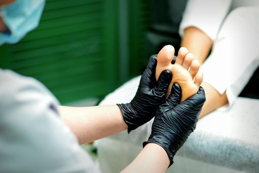 Beautician in protective rubber gloves doing massage on the sole of the female foot in a spa beauty salon