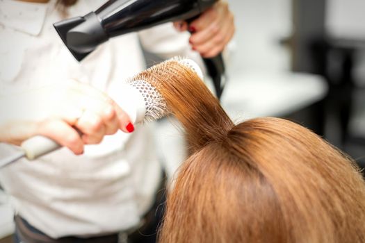 Close up of hairdresser drying long red hair with a hairdryer and round brush in a beauty salon