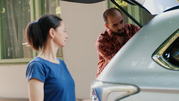 Parents putting voyage luggage and baggage inside car trunk while going on summer holiday field trip. Casual family going on summer cityscape tour while loading trolleys inside vehicle.