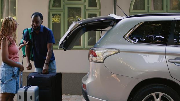 African american couple loading voyage luggage in car while getting ready for summer citybreak departure. Boyfriend and girlfriend putting baggage and trolleys in vehicle trunk.