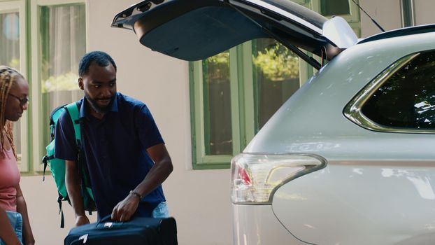 Positive african american couple putting voyage luggage inside car trunk while getting ready for holiday summer citybreak. Casual people loading vehicle with trolleys while going on field trip.