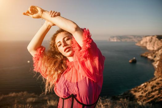 Close up shot of beautiful young caucasian woman with curly blond hair and freckles looking at camera and smiling. Cute woman portrait in a pink long dress posing on a volcanic rock high above the sea