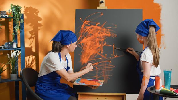 Mom and child learning to paint with orange aquarelle color and paintbrush, creating artwork design together on canvas. Happy people painting masterpiece with watercolor and brush.