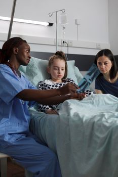 African american nurse showing radiography scan image to sick girl while mother sitting beside her. Pediatric healthcare facility staff analyzing MRI results of ill kid under treatment resting