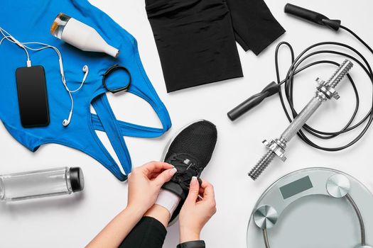 Young woman in sporting leggings laces sneakers, preparing for training. Accessories for sports, bottle of water, smartphone, headphones, fitness bracelet, dumbbells on white background flat lay top view. Fitness concept