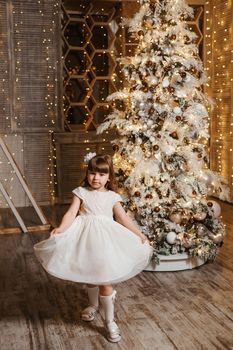 A girl in a festive light dress next to a Christmas tree, lights of garlands in the background. The concept of New Year holidays.
