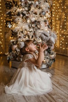 A girl in a festive light dress next to a Christmas tree, lights of garlands in the background. The concept of New Year holidays.