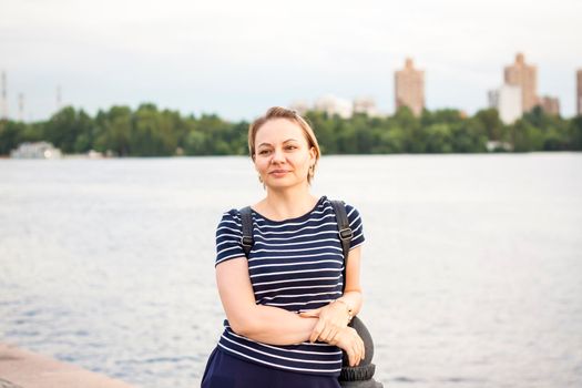 A girl in a striped jacket is standing on the river embankment, leaning on the parapet, looking into the distance. Evening walk around the city