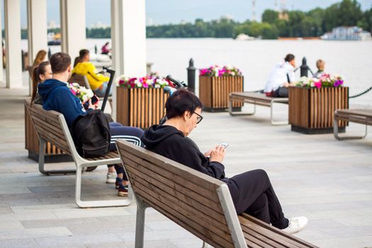 2022-07-17. Russia. Moscow. Northern River Station. A woman is sitting on a bench with a phone in her hands on the embankment. Other people are resting nearby. Moscow River