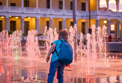 Children have fun frolicking in the fountains near the building in the park in the sunset light. Evening walk around the city