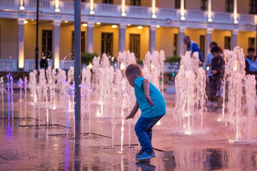 Children have fun frolicking in the fountains near the building in the park in the sunset light. Evening walk around the city