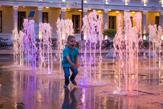 Children have fun frolicking in the fountains near the building in the park in the sunset light. Evening walk around the city