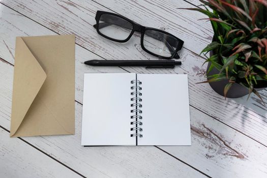 Notepad with brown envelop, reading glasses, pen and potted plant on wooden desk. Flat lay. Copy space.
