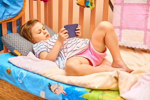 a young human being below the age of puberty or below the legal age of majority. A cute child lying in a crib looking news, games, fairy tales on a smartphone.