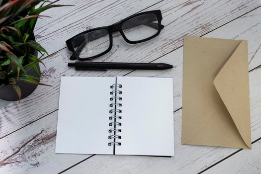 Notepad with brown envelop, reading glasses, pen and potted plant on wooden desk. Flat lay. Copy space.