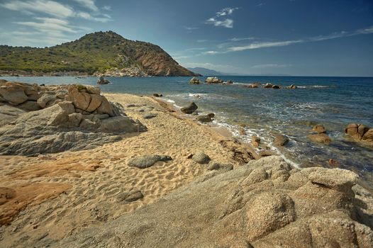 Beautiful seascape of the Cala Sa Figu beach in the south of Sardinia, with the rocks in the foreground, the beach and the curly mountains of vegetation in the background during the summer.