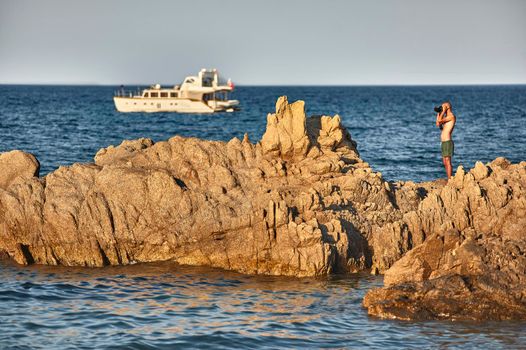 Photographer intent on photographing a natural landscape standing on top of a cliff in the middle of the blue sea during sunset.