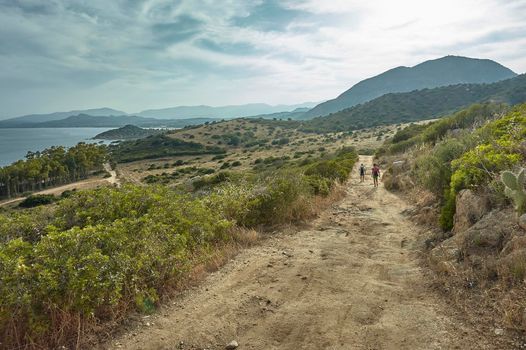 Couple of hikers walking in a mountain dirt road on the southern Mediterranean coast of Sardinia surrounded by a breathtaking natural landscape.