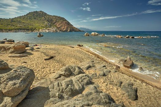 Beautiful seascape of the Cala Sa Figu beach in the south of Sardinia, with the rocks in the foreground, the beach and the curly mountains of vegetation in the background during the summer.