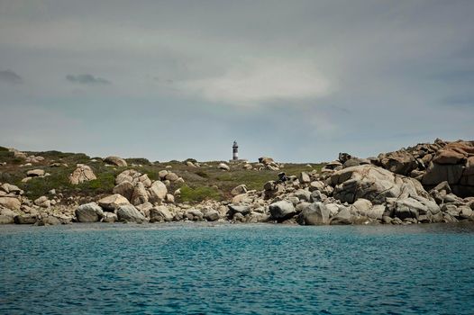 Lighthouse in the middle of the rocky landscape of the southern coast of Sardinia taken from the sea on a cloudy and gloomy day.