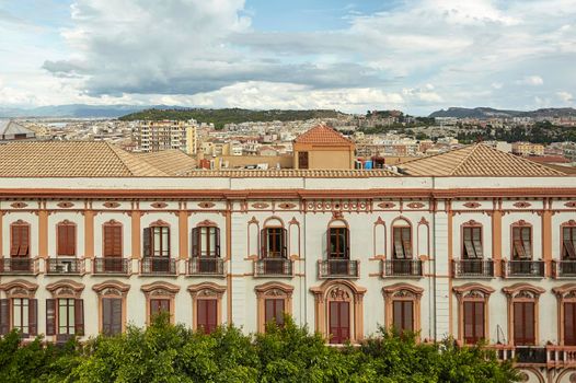 Side detail of a historic building in the city center of Cagliari in Italy, with behind the city panorama under a sky full of clouds.