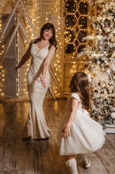 A little girl with her mother in light festive dresses next to the Christmas tree. The theme of New Year's holidays and festive interior with garlands and light bulbs.