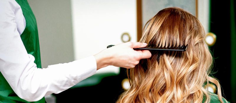 Back view of hairdresser combing wavy hair of a young blonde woman in a beauty salon