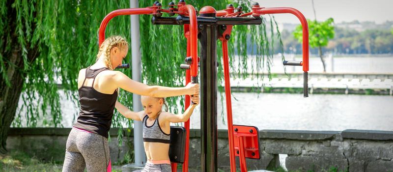 Mother with daughter in sportswear is doing sport exercises on a simulator in the summer park