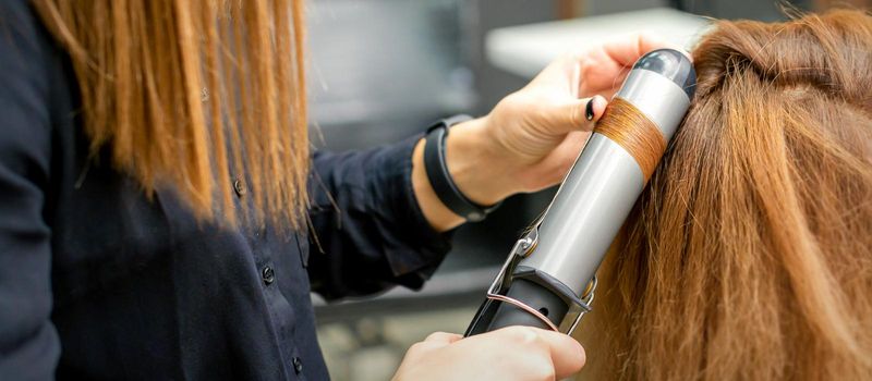 Professional hairdresser makes curls with a curling iron for a young woman with long red hair in a beauty salon